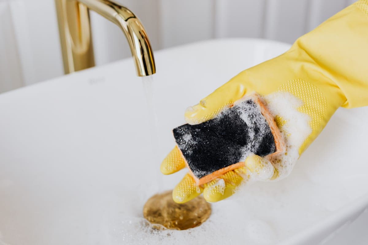 A gloved hand cleaning a white sink with a sponge and detergent